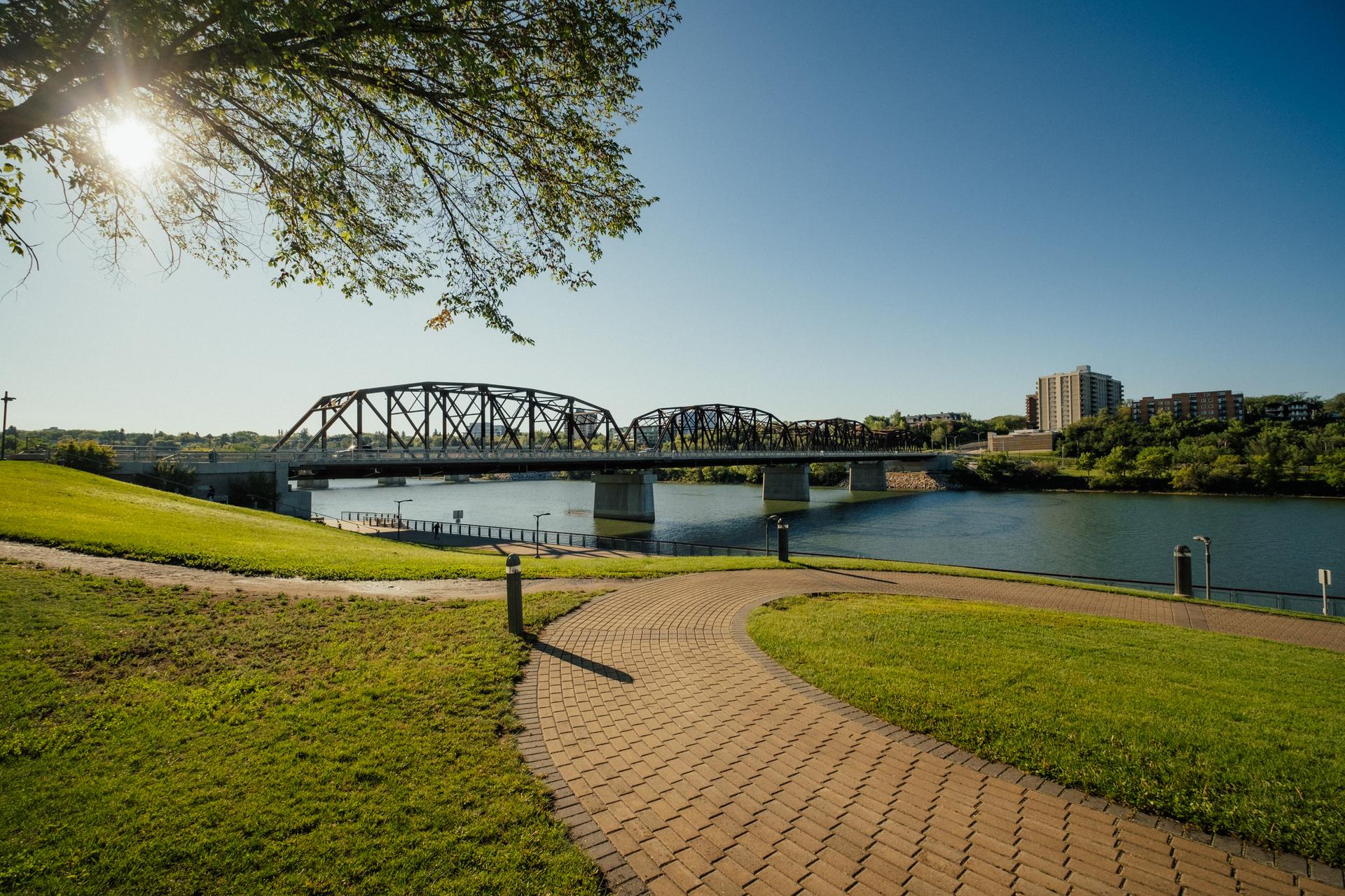 A pedestrian stone pathway leading up to a bridge crossing a river on a sunny day.