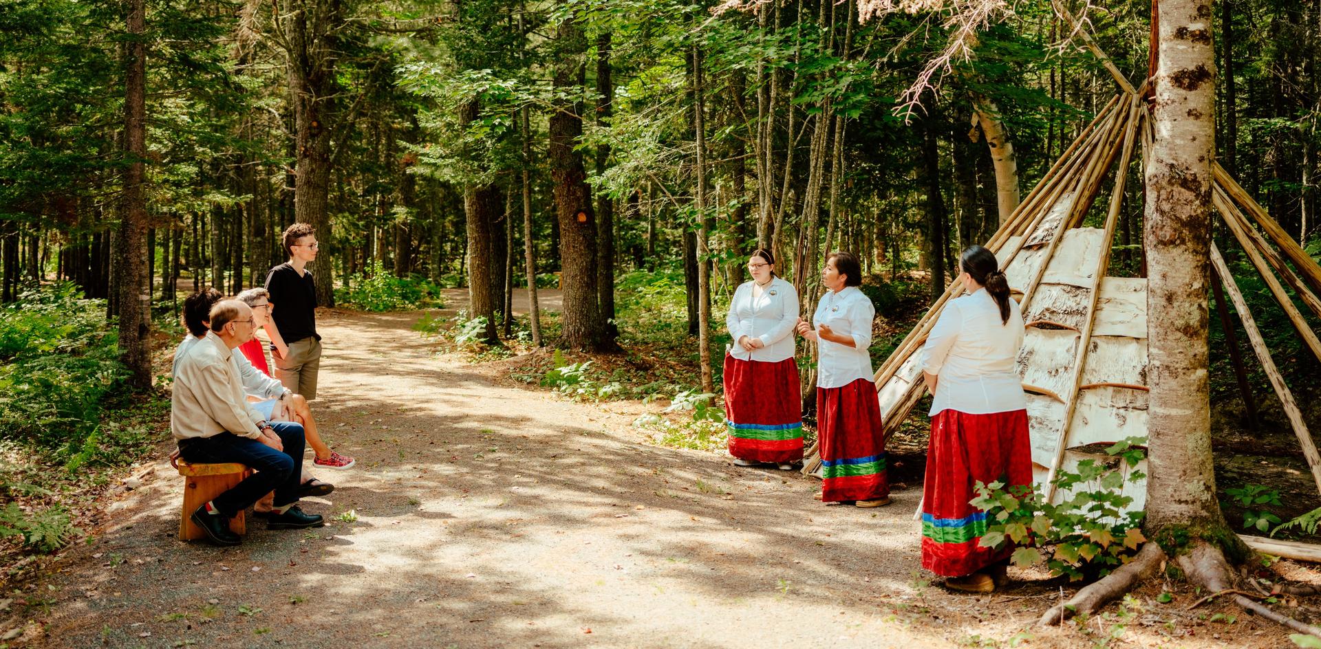 A group of visitors listens to three Indigenous people in traditional attire in a forest setting
