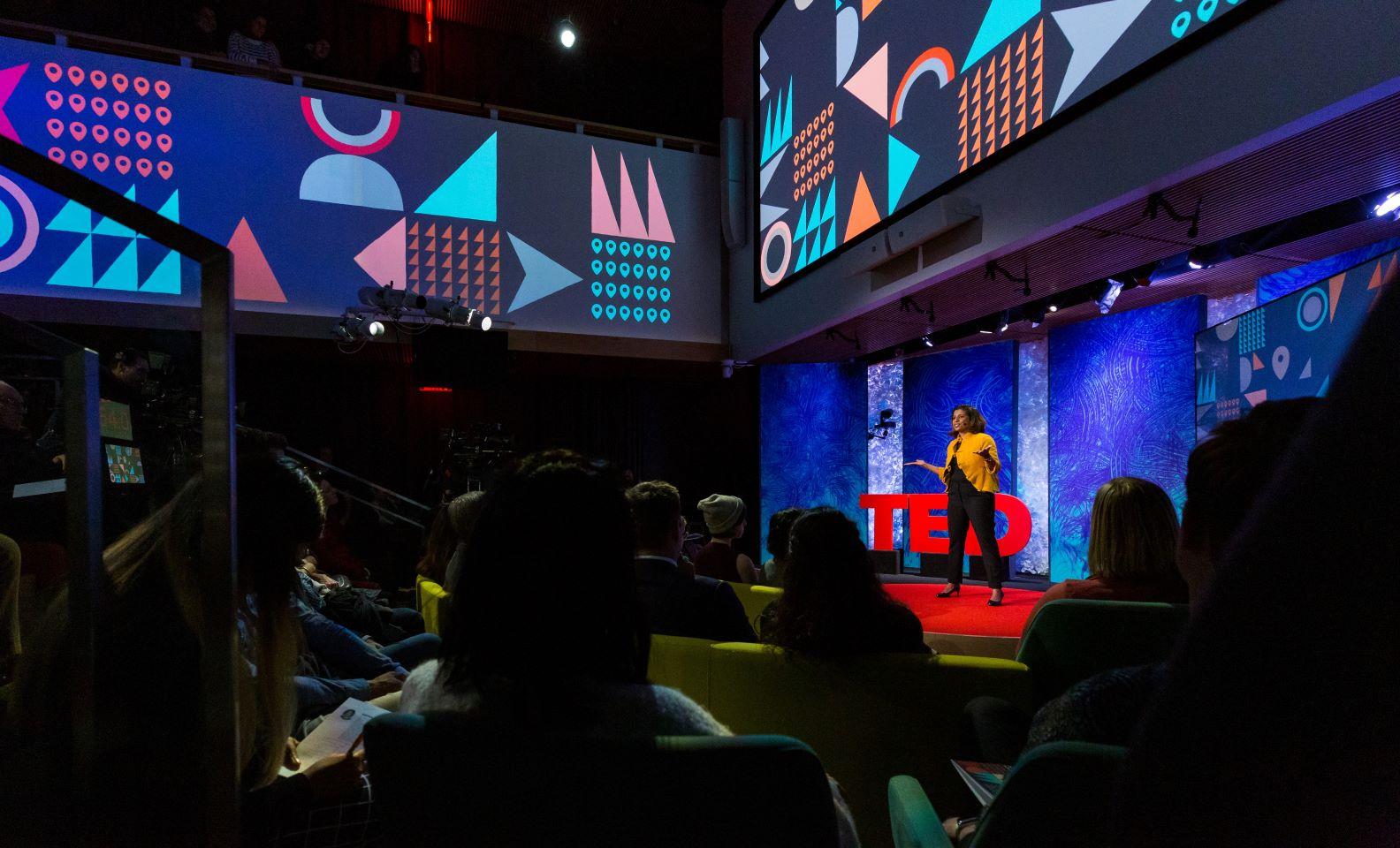 A woman stands on a TED talk stage giving a speech to an audience.