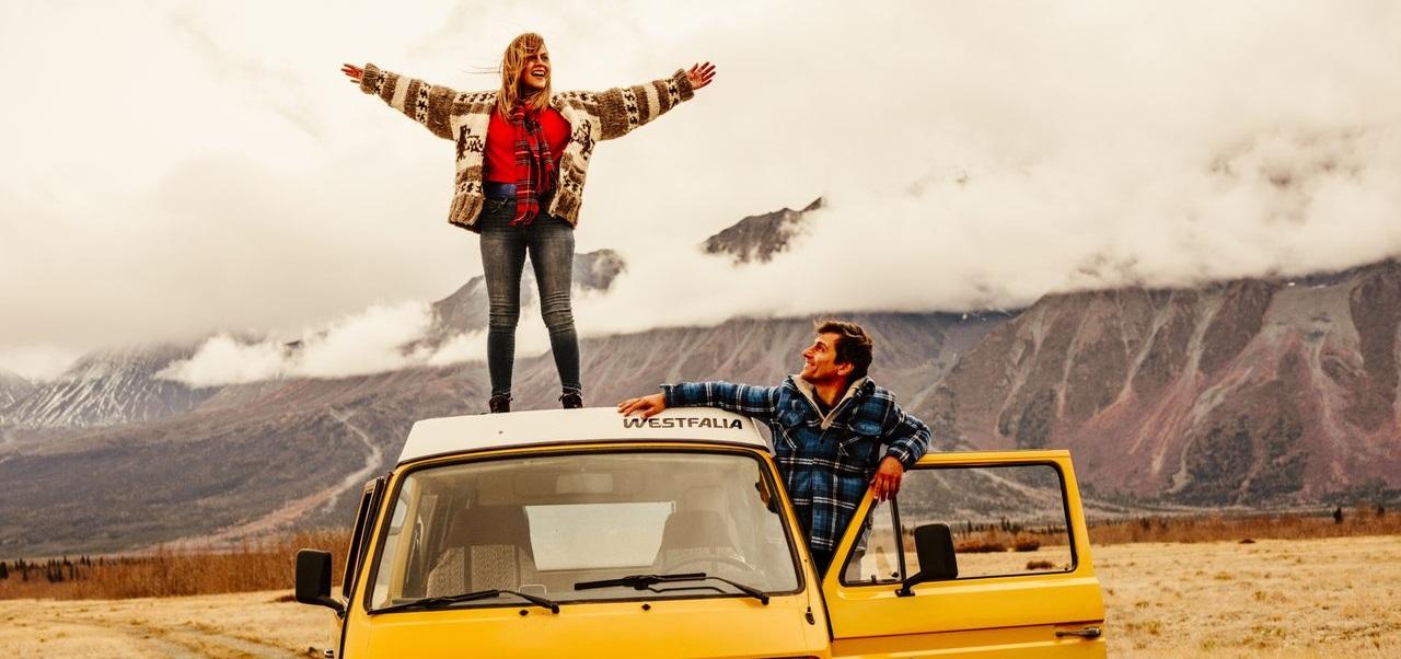 A woman stands on the roof of a yellow VW Westalia bus with her arms out stretched and a smile on her face. A man leaning out the driver's side window looking up at her.