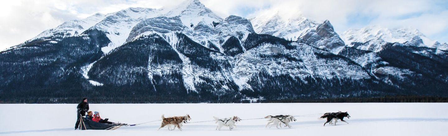 Sled dogs pull a sled through a snowy valley. Snow capped mountains are in the background.