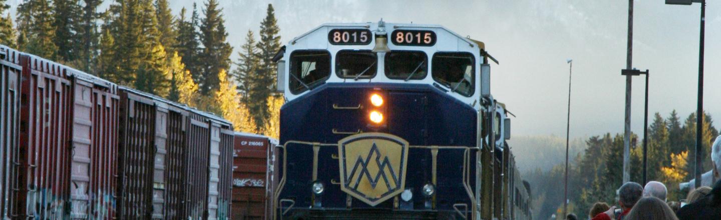 A train with a yellow mountain logo on the front sits still on track while people wait to board on the side platform. Behind the train stands a tall white mountain.