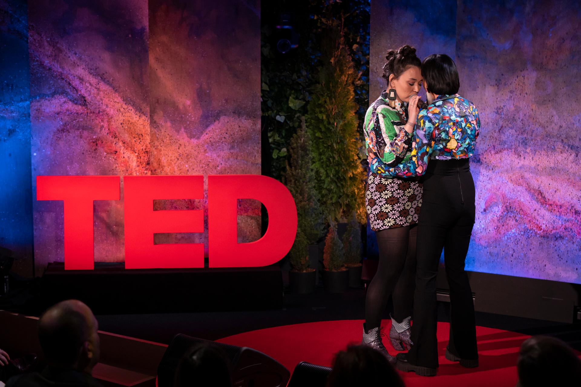 Two indigenous women stand facing each other on a TED talk stage with microphones.