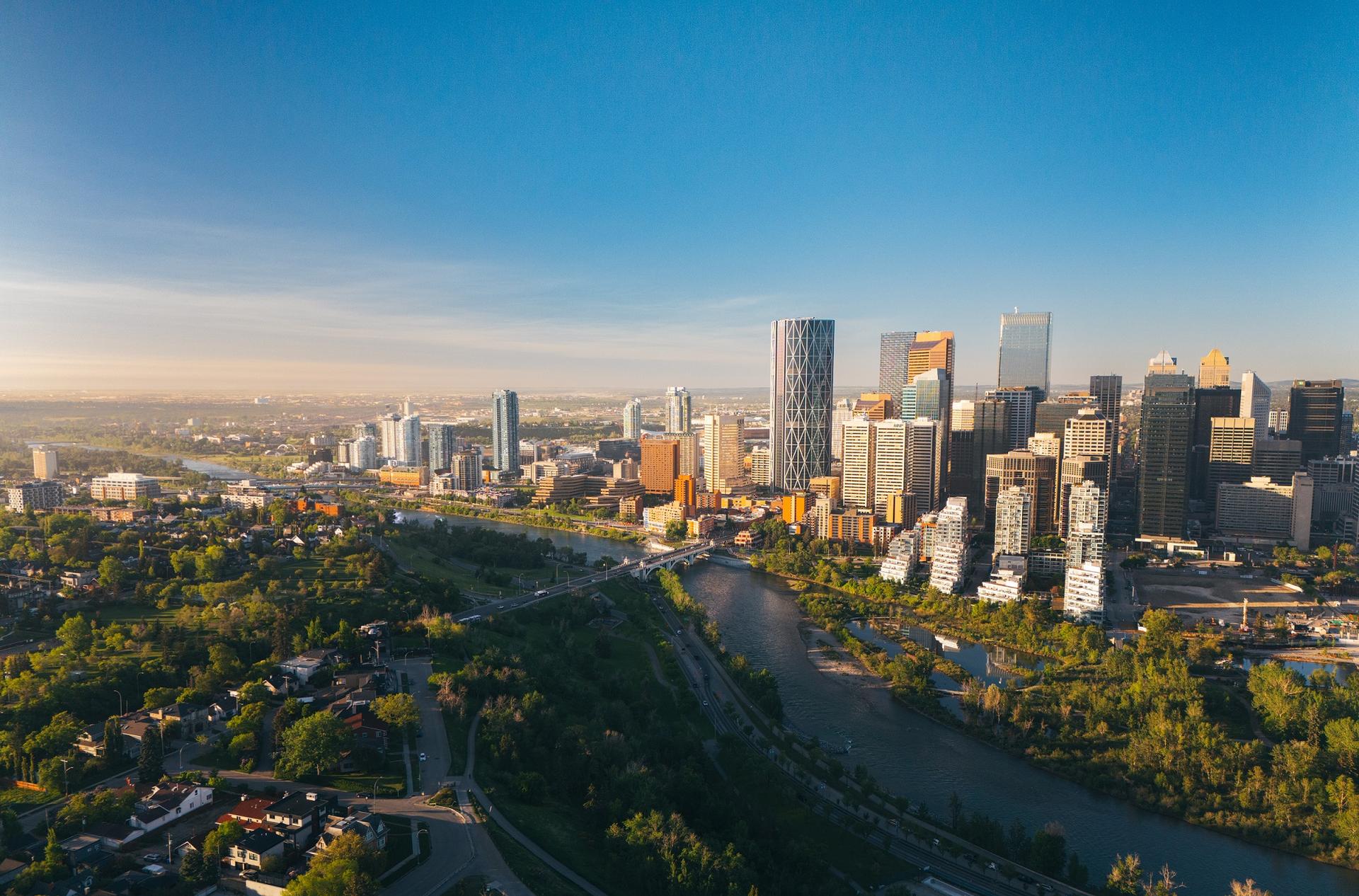 Calgary skyline at golden hour, with modern downtown towers rising above the Bow River and lush green parkland under a clear blue sky