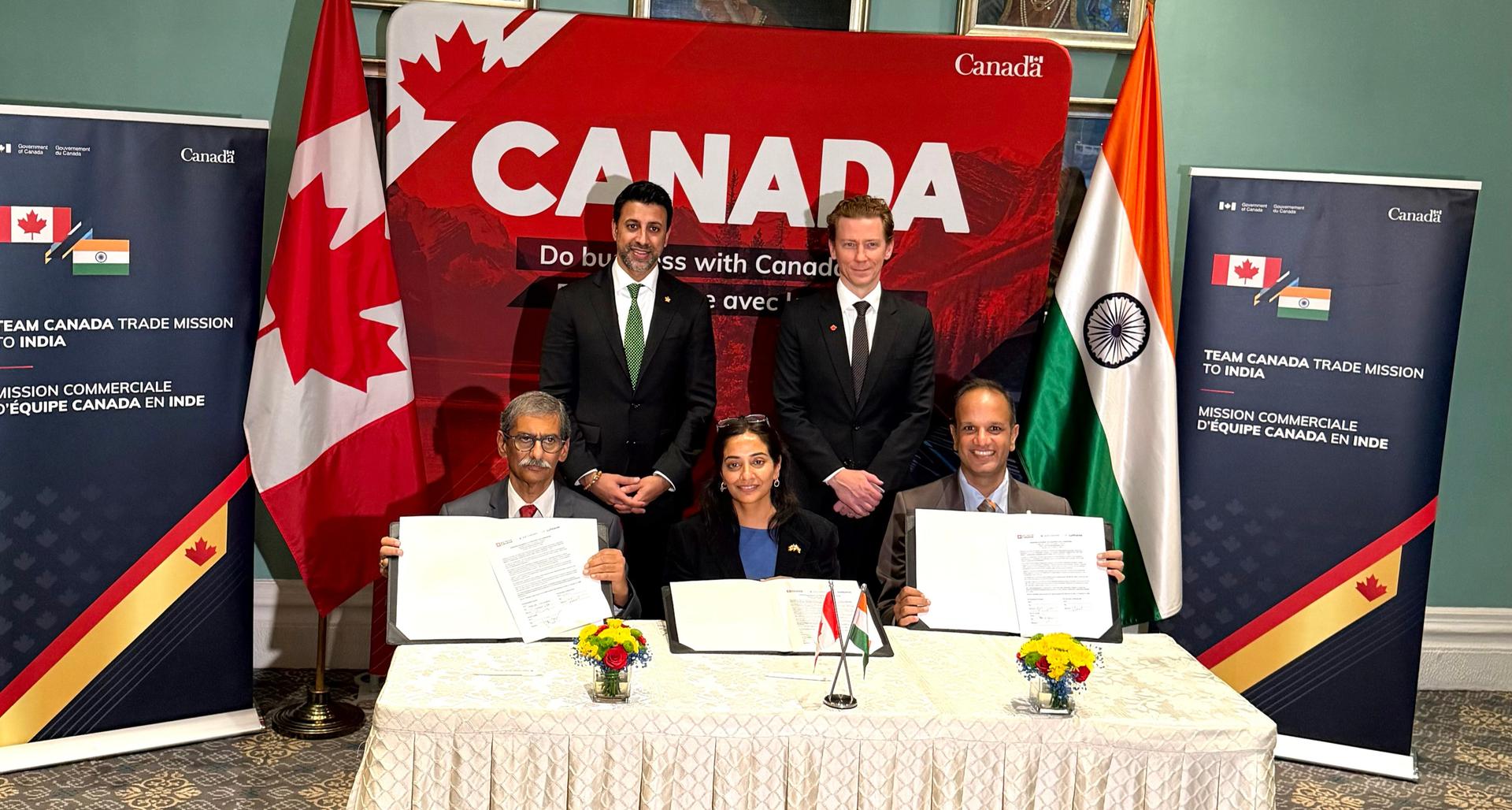 Three seated representatives display signed documents while two officials stand behind them, with Canadian and Indian flags and Team Canada Trade Mission to India banners in the background.
