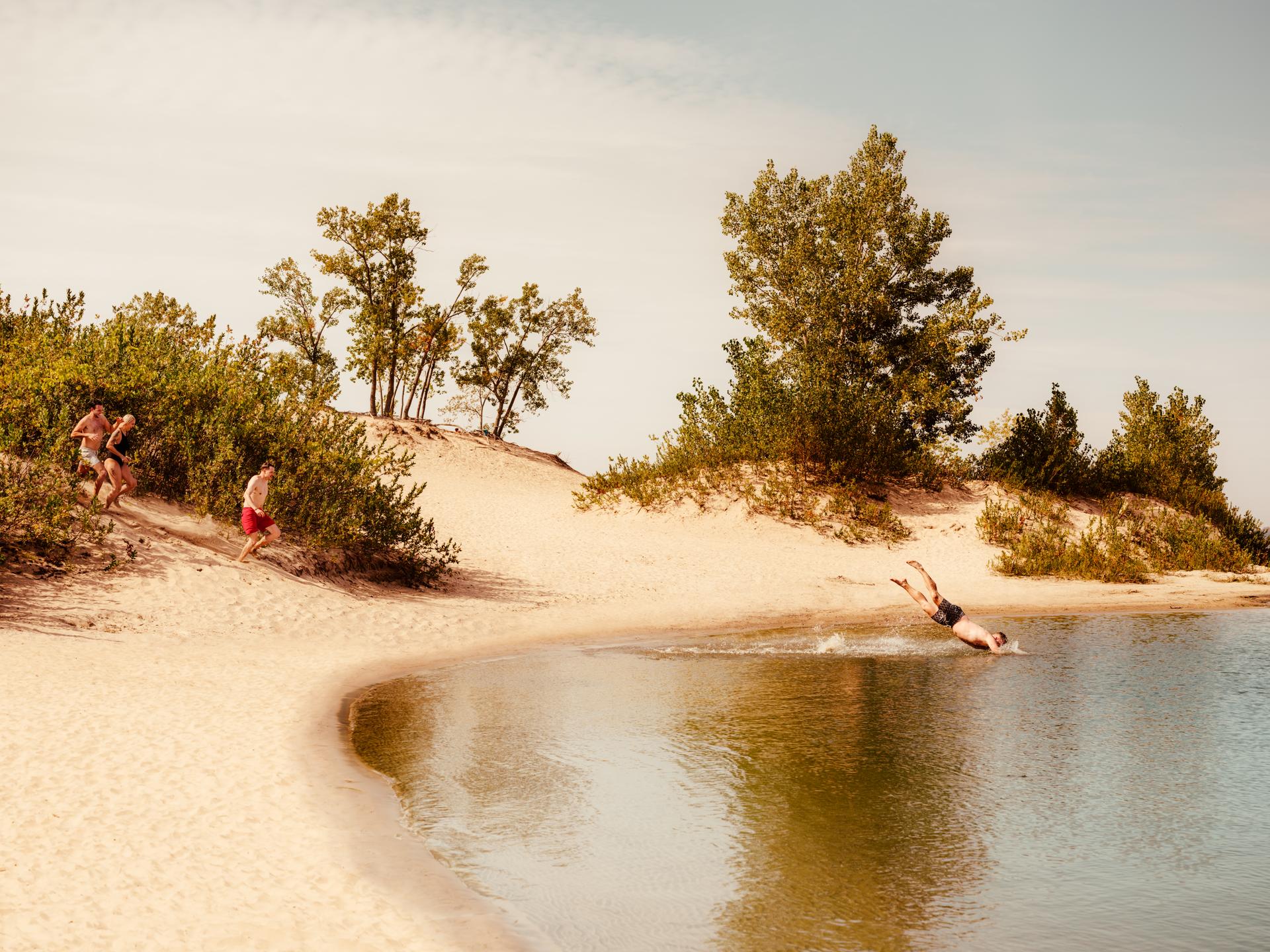 Trois personnes en maillot de bain courent sur une dune de sable aux abords d’un lac. Une quatrième personne, à l'avant, plonge dans l'eau. Le sable autour du lac est bordé d'arbres et d'arbustes verts.Trois personnes en maillot de bain courent sur une dune de sable aux abords d’un lac. Une quatrième personne, à l'avant, plonge dans l'eau. Le sable autour du lac est bordé d'arbres et d'arbustes verts.